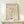 May include: Framed beige poster with black line drawing of woman's face holding a dove, displayed on light wood shelf against cream wall. Simple continuous line art style showing feminine features and bird. Small wrapped gift box with greenery sits beside the natural wood frame.