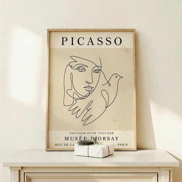 May include: Framed beige poster with black line drawing of woman's face holding a dove, displayed on light wood shelf against cream wall. Simple continuous line art style showing feminine features and bird. Small wrapped gift box with greenery sits beside the natural wood frame.
