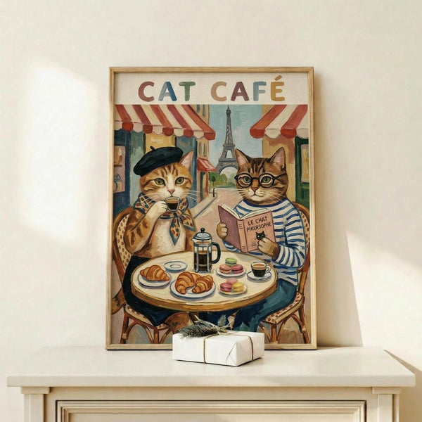 May include: Framed vintage-style poster showing two anthropomorphic cats at a Parisian cafe table. One orange tabby wears a black beret and patterned scarf while sipping coffee, the other gray tabby wears glasses and striped shirt while reading. The Eiffel Tower appears in the background between red-striped awnings. The table holds croissants, macarons, and a French press. Light wood frame sits on white shelf next to small wrapped gift.