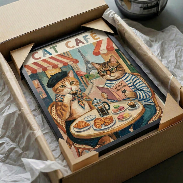 May include: Framed vintage-style poster featuring two anthropomorphic cats at a Parisian cafe table. One cat wears a black beret and striped shirt, the other reads a book wearing glasses and striped top. The scene shows croissants, coffee, macarons, and the Eiffel Tower in the background. The artwork is in a black frame, nestled in white tissue paper inside a brown cardboard shipping box.