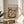 May include: Framed poster artwork displayed on a wooden side table next to a beige upholstered chair. The illustration shows two cats in French cafe setting - one orange tabby wearing a black beret holding coffee, another tabby with glasses in striped shirt reading. Background features Parisian storefronts with red striped awnings and Eiffel Tower. Table holds croissants, coffee cups, and French press. Warm sunlight streams across the neutral-toned room with plant visible in corner.