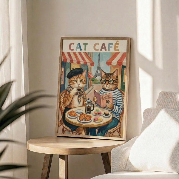 May include: Framed poster artwork displayed on a wooden side table next to a beige upholstered chair. The illustration shows two cats in French cafe setting - one orange tabby wearing a black beret holding coffee, another tabby with glasses in striped shirt reading. Background features Parisian storefronts with red striped awnings and Eiffel Tower. Table holds croissants, coffee cups, and French press. Warm sunlight streams across the neutral-toned room with plant visible in corner.
