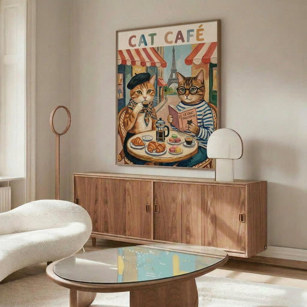 May include: Framed poster showing two cats at a Parisian cafe table with the Eiffel Tower in background. One orange tabby wears a black beret and holds coffee, the other gray tabby in striped shirt reads a French newspaper. Colorful vintage-style artwork with red cafe awnings. Displayed on wooden credenza with white curved chair and glass coffee table in modern living room setting.