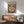 May include: Framed vintage-style poster showing two anthropomorphic cats at a Parisian sidewalk cafe, one orange tabby in beret and one striped cat reading, with Eiffel Tower backdrop. Displayed above mid-century modern wood credenza with white ceramic lamp, next to sage green upholstered armchair in bright, naturally-lit room with white walls.