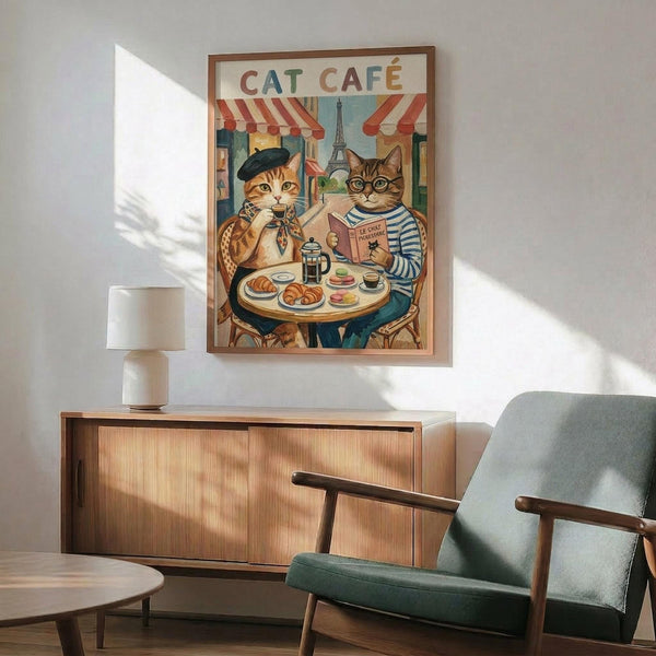 May include: Framed vintage-style poster showing two anthropomorphic cats at a Parisian sidewalk cafe, one orange tabby in beret and one striped cat reading, with Eiffel Tower backdrop. Displayed above mid-century modern wood credenza with white ceramic lamp, next to sage green upholstered armchair in bright, naturally-lit room with white walls.