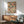 May include: A framed vintage-style poster featuring a colorful mosaic pattern of stacked books in various sizes and colors including blues, reds, oranges, and creams. The artwork hangs above a mid-century modern wooden credenza with sliding doors and warm wood grain. A minimalist white ceramic table lamp sits on the credenza next to a sage green upholstered armchair with wooden arms. Natural sunlight creates geometric shadows on the white wall, and a round wooden coffee table is partially visib
