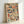 A framed art print featuring densely packed book spines arranged in a mosaic pattern, displayed in various colors including navy blue, rust orange, teal, cream, burgundy, and forest green. The books appear stacked both horizontally and vertically creating a textured, layered library aesthetic. The artwork is housed in a natural wood frame and leans against a white wall, casting a soft shadow.