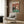 May include: Whimsical poster featuring two cats in formal attire playing poker at a green casino table with cards, chips, and wine. The tabby cat wears a black tuxedo while the orange cat sports an elegant dress. Red fleur-de-lis wallpaper background. Displayed above a cream upholstered chair with geometric pillow in a modern living room with wood paneling, brass floor lamp, and side table.