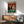 May include: Framed vintage-style poster showing two cats in formal attire playing poker at a green casino table with chips, cards, and wine. The tabby cat wears a black tuxedo while the orange cat sports an elegant dress and holds cards. Red damask wallpaper background. Displayed above a wooden sideboard with lamp in modern living room setting with mint green armchair.