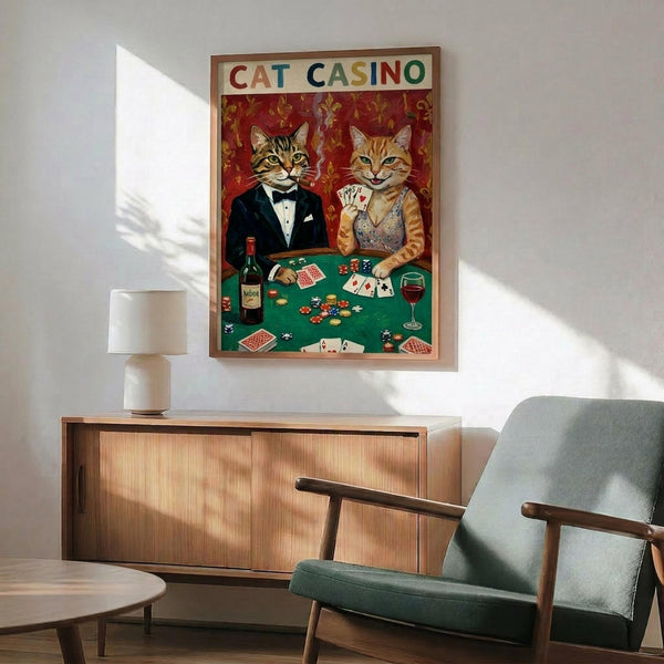 May include: Framed vintage-style poster showing two cats in formal attire playing poker at a green casino table with chips, cards, and wine. The tabby cat wears a black tuxedo while the orange cat sports an elegant dress and holds cards. Red damask wallpaper background. Displayed above a wooden sideboard with lamp in modern living room setting with mint green armchair.