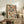 May include: Framed artwork displaying a colorful mosaic pattern of book spines in various sizes, featuring blues, oranges, reds, and cream tones. The piece sits on a rustic wooden sideboard alongside a potted fern, ceramic vase, and stacked books. A beige upholstered chair with brown throw pillows is positioned nearby against a light neutral wall.