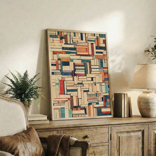 May include: Framed artwork displaying a colorful mosaic pattern of book spines in various sizes, featuring blues, oranges, reds, and cream tones. The piece sits on a rustic wooden sideboard alongside a potted fern, ceramic vase, and stacked books. A beige upholstered chair with brown throw pillows is positioned nearby against a light neutral wall.
