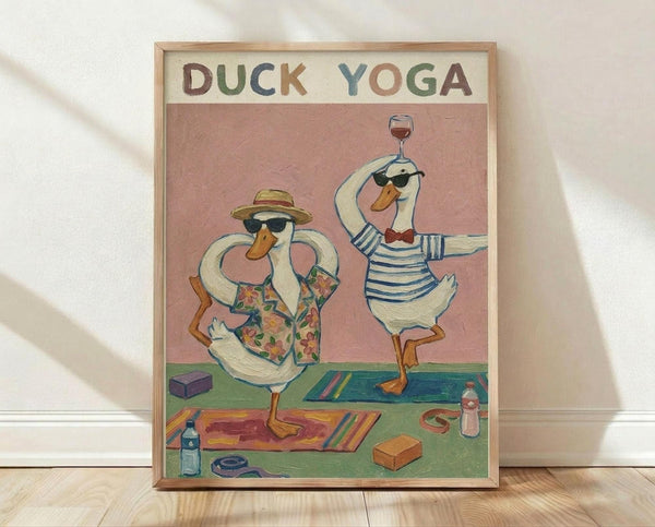 May include: Framed poster showing two cartoon ducks in sunglasses doing yoga poses on colorful mats. One duck wears a floral shirt and sun hat in tree pose, the other in striped shirt balances with wine glass. Pink background with yoga props scattered around including blocks, water bottle, and towel. Light wood frame against white wall on wooden floor.