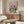 May include: Framed poster showing two cats wearing sunglasses having a picnic with wine, cheese, and grapes under a pink sky, displayed above a mid-century modern walnut credenza in a minimalist living room with white upholstered chair and glass coffee table