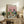 May include: Framed poster print displayed on rustic wooden credenza in cozy living room setting. Artwork features two cats wearing sunglasses at outdoor picnic with wine glasses, cheese, grapes, and bread on checkered blanket under pink sky with clouds. Colorful vintage-style illustration in natural wood frame, surrounded by fern plant, books, ceramic vase, and cream armchair.