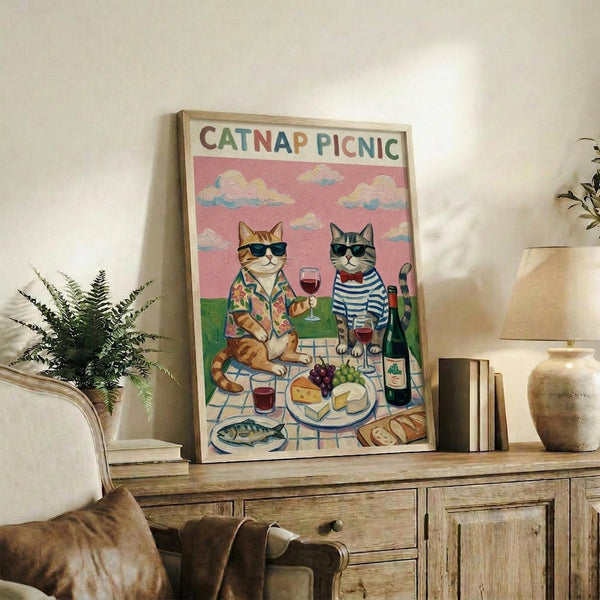 May include: Framed poster print displayed on rustic wooden credenza in cozy living room setting. Artwork features two cats wearing sunglasses at outdoor picnic with wine glasses, cheese, grapes, and bread on checkered blanket under pink sky with clouds. Colorful vintage-style illustration in natural wood frame, surrounded by fern plant, books, ceramic vase, and cream armchair.