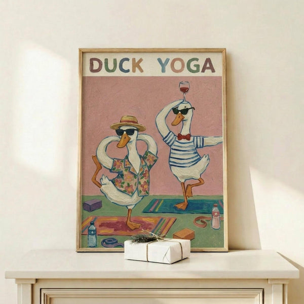 May include: Framed vintage-style poster showing two white ducks in sunglasses doing yoga poses on colorful mats. One duck wears a straw hat and floral shirt, the other a striped shirt while balancing a wine glass. Pink background with yoga props scattered around including blocks, water bottles, and towels. Light wood frame displayed on white wooden surface next to small wrapped gift with evergreen sprig.