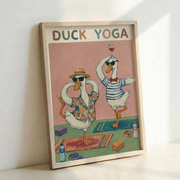 Framed poster featuring two cartoon ducks doing yoga poses on colorful mats. One duck wears a Hawaiian shirt and sun hat while balancing on one leg, the other wears a striped shirt and holds a wine glass while standing on one foot. The illustration has a pink background with "DUCK YOGA" text at top in multicolored letters. Yoga props like blocks and water bottles are scattered around the green grass setting. The artwork is displayed in a light wooden frame leaning against a white wall.