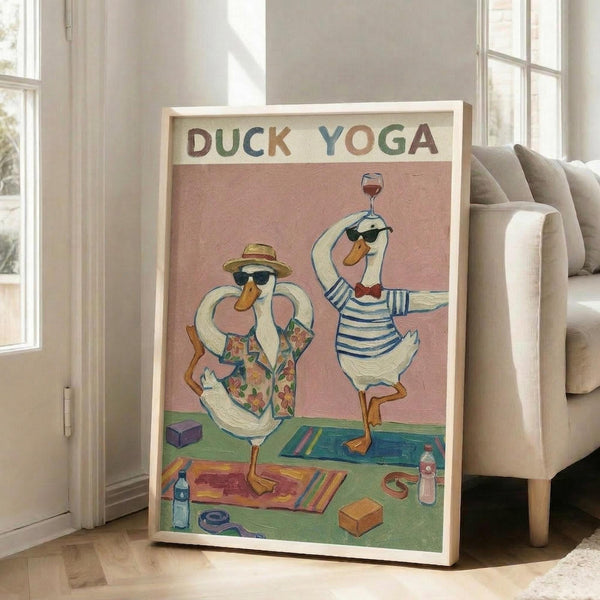 May include: Framed poster showing two white ducks in sunglasses doing yoga poses on colorful mats. One duck wears a straw hat and floral shirt, the other a striped shirt while balancing a wine glass. Pink background with "DUCK YOGA" text in multicolored letters. Yoga props scattered on green floor. Light wood frame leaning against beige chair in bright, modern living room with large windows.