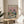 May include: Framed poster print on wooden side table next to beige upholstered chair and leafy plant. Artwork shows two cats wearing sunglasses at outdoor picnic - orange tabby in floral shirt holding wine glass, gray striped cat in blue striped shirt. Picnic spread includes wine bottle, cheese, grapes, fish, and bread on checkered blanket. Pink sky with fluffy clouds background. Colorful "CATNAP PICNIC" text at top. Natural lighting creates soft shadows in neutral-toned room setting.