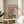 May include: Framed whimsical illustration showing two white ducks wearing sunglasses doing yoga poses on colorful mats against a pink background. One duck wears a floral shirt and sun hat, the other a striped shirt while balancing a wine glass. Yoga props like blocks and water bottles scattered around. The wooden-framed poster sits on a light wood round side table next to a beige upholstered chair, with natural sunlight streaming across white walls and plant leaves visible.