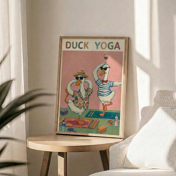 May include: Framed whimsical illustration showing two white ducks wearing sunglasses doing yoga poses on colorful mats against a pink background. One duck wears a floral shirt and sun hat, the other a striped shirt while balancing a wine glass. Yoga props like blocks and water bottles scattered around. The wooden-framed poster sits on a light wood round side table next to a beige upholstered chair, with natural sunlight streaming across white walls and plant leaves visible.