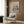 May include: Whimsical poster featuring two cats wearing sunglasses at a picnic with wine, cheese, and bread under a pink cloudy sky, displayed in light wooden frame on wood-paneled wall above beige upholstered chair with geometric pillow, brass floor lamp and side table with open book nearby