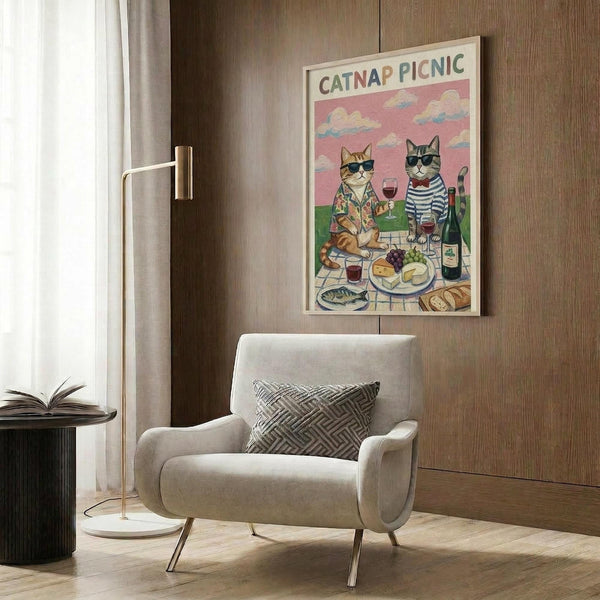 May include: Whimsical poster featuring two cats wearing sunglasses at a picnic with wine, cheese, and bread under a pink cloudy sky, displayed in light wooden frame on wood-paneled wall above beige upholstered chair with geometric pillow, brass floor lamp and side table with open book nearby