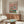 May include: Framed whimsical illustration of two cartoon ducks doing yoga poses on colorful mats, one wearing a floral shirt and sun hat, the other in striped shirt holding a wine glass, displayed above a mid-century modern walnut credenza with white mushroom lamp in a minimalist living room with cream walls, white upholstered chair, and glass-top oval coffee table.