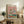 May include: Framed whimsical poster showing two cartoon ducks in sunglasses doing yoga poses on colorful mats, one wearing a floral shirt and hat, the other in striped shirt holding wine glass, displayed on rustic wooden sideboard with plant, books, ceramic vase, and beige armchair in cozy living room setting with white walls