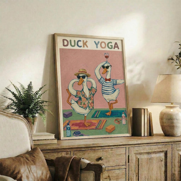 May include: Framed whimsical poster showing two cartoon ducks in sunglasses doing yoga poses on colorful mats, one wearing a floral shirt and hat, the other in striped shirt holding wine glass, displayed on rustic wooden sideboard with plant, books, ceramic vase, and beige armchair in cozy living room setting with white walls