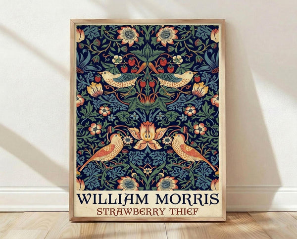 May include: Framed poster print featuring William Morris's ornate Victorian pattern with symmetrical design of birds, strawberry plants, and flowers in rich blues, greens, and coral tones on dark navy background. Light wood frame leans against white wall on light wooden floor with soft natural lighting creating gentle shadows.