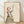 May include: Framed art print leaning against white wall on light wood floor. Artwork shows stylized woman silhouette filled with layered landscape scenes including forests, mountains, rivers, and small cabins in earthy greens, blues, and browns. Woman has flowing red hair and reaches toward mountain outline with small golden tree. Natural wood frame, cream background, soft natural lighting creates shadows on textured white wall.