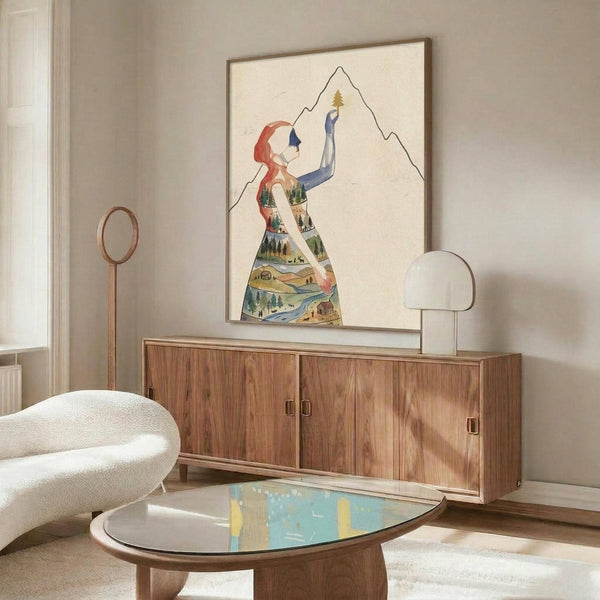 May include: Framed poster artwork displayed above wooden sideboard in modern living room setting. Artwork shows stylized woman figure with red hair reaching upward toward mountain peak, her dress filled with colorful landscape scenes including trees, water, and small buildings. Warm wood furniture, white curved chair, glass coffee table, and white table lamp complete the mid-century modern room scene.