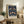 May include: Framed poster with ornate blue floral pattern featuring symmetrical birds, flowers, and foliage in rich navy, green, coral, and cream colors displayed on rustic wooden sideboard beside fern plant, ceramic vases, and books in cozy living room with beige upholstered chair and neutral curtains