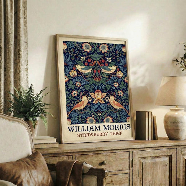 May include: Framed poster with ornate blue floral pattern featuring symmetrical birds, flowers, and foliage in rich navy, green, coral, and cream colors displayed on rustic wooden sideboard beside fern plant, ceramic vases, and books in cozy living room with beige upholstered chair and neutral curtains