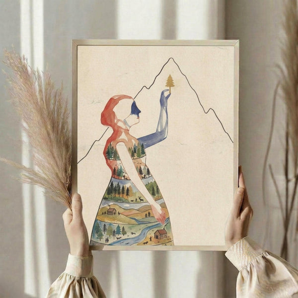May include: Person holding framed poster showing silhouette of woman with red hair reaching toward mountain peak with small tree. Woman's body filled with layered landscape scenes including forests, cabins, rivers, and rolling hills in watercolor style. Light wood frame, neutral cream background with dried pampas grass visible.
