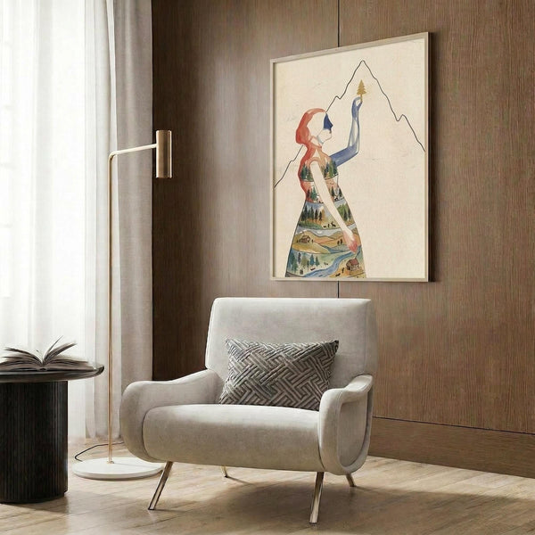 May include: Contemporary living room scene featuring a framed art print on wood-paneled wall above beige upholstered armchair with geometric pillow. The artwork shows a stylized female silhouette filled with colorful landscape elements including mountains, trees, and water, rendered in watercolor style with red hair detail. Modern brass floor lamp and dark round side table with open book complete the minimalist interior setting.