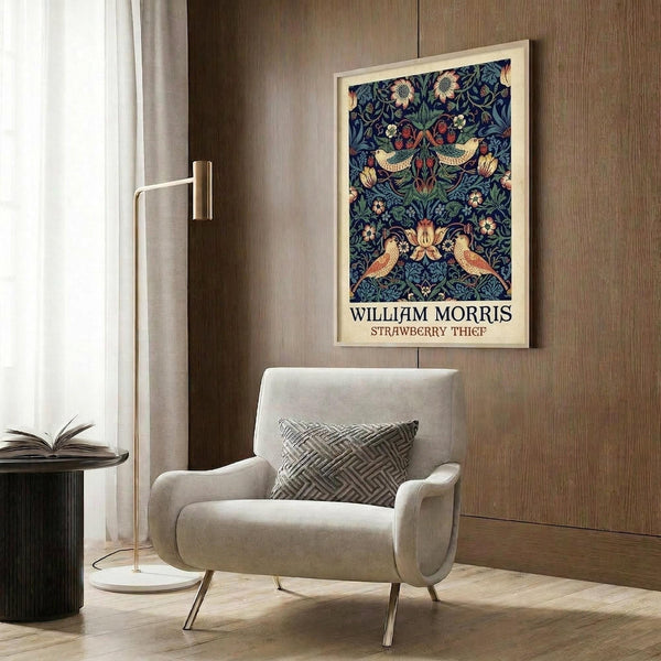 May include: Framed William Morris Strawberry Thief poster with intricate blue floral pattern featuring birds and flowers, displayed on wood-paneled wall above beige upholstered armchair with geometric pillow. Modern brass floor lamp and dark round side table with open book complete the warm, contemporary reading nook setting.
