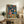 May include: Framed poster featuring a cartoon cat in pink spacesuit and golden retriever in blue spacesuit sharing fried chicken and drinks in space, displayed on rustic wooden console table with books, ceramic vases, fern plant, and beige sofa in modern living room setting
