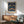 May include: Framed William Morris Strawberry Thief poster displayed above a wooden credenza in a mid-century modern living room. The vintage-style print features symmetrical birds, strawberries, and floral patterns in rich blues, greens, and coral tones against a dark background. A sage green upholstered chair with wooden arms sits beside the credenza, along with a small white table lamp and round coffee table.