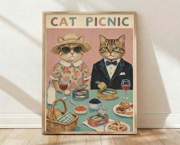 May include: Framed vintage-style poster showing two dressed-up cats at a picnic table. Left cat wears floral dress, sun hat and sunglasses; right cat in navy suit with bow tie. Table displays wine glasses, caviar bowls, canned fish, pastries and wicker basket. Warm pink and mint green color palette with cream background. Light wooden frame against white wall on wooden floor with soft shadows.