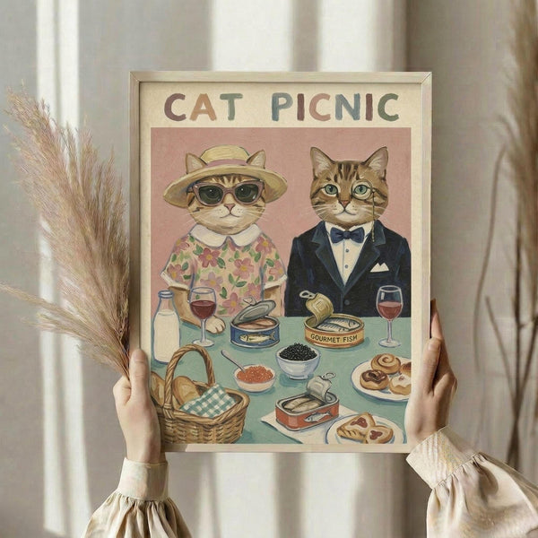 May include: Person holding framed vintage-style illustration showing two dressed cats at picnic table with gourmet food including caviar, wine, and canned fish. Female cat wears floral dress and sun hat, male cat in navy suit with bow tie. Warm beige tones with pink background. Person also holds dried wheat stems against neutral curtained backdrop.