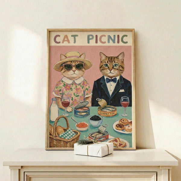 May include: Framed vintage-style illustration showing two anthropomorphic cats at a picnic table. Left cat wears floral dress, sun hat and sunglasses; right cat sports navy suit with bow tie. Table displays wine glasses, caviar, canned fish, pastries and picnic basket against pink background. Cream-colored frame sits on light wood console with small wrapped gift nearby on white wall.