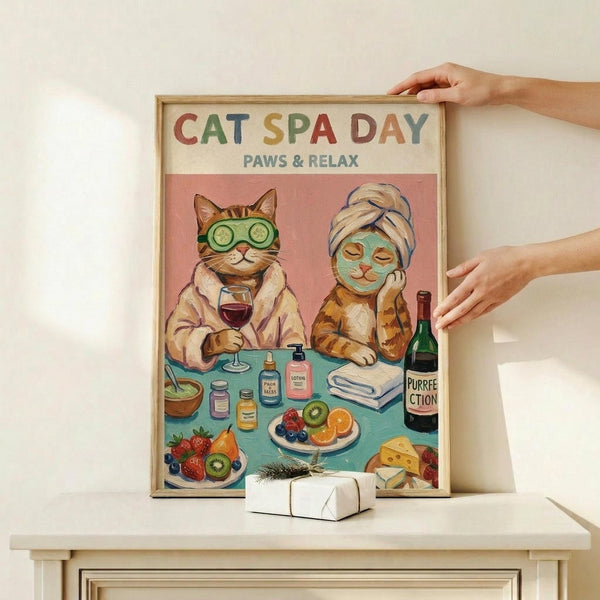 May include: Framed poster with wooden frame showing two illustrated cats at spa day - one wearing cucumber eye mask and towel head wrap, another with green face mask holding wine glass. Colorful artwork features spa items, fruits, and "PURRFECTION" wine bottle on turquoise table. Hands positioning frame on white wooden shelf with small wrapped gift below.
