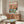 May include: Framed poster print displayed above wooden sideboard in modern living room. Two cats at spa table - orange tabby wearing cucumber eye mask and robe holding wine glass, orange cat with face mask and head wrap. Table has colorful fruits, spa products, wine bottle labeled "PURRFECTION". White curved chair and glass coffee table in foreground. Warm wood tones and neutral walls create cozy interior setting.