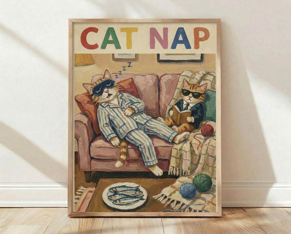 May include: Framed poster print leaning against white wall on light wooden floor. Artwork shows two orange tabby cats on pink sofa - one sleeping in striped blue pajamas with eye mask, another reading while wearing suit and sunglasses. Colorful "CAT NAP" text at top. Scene includes yarn balls, fish plate, and cozy home details in warm illustrated style.
