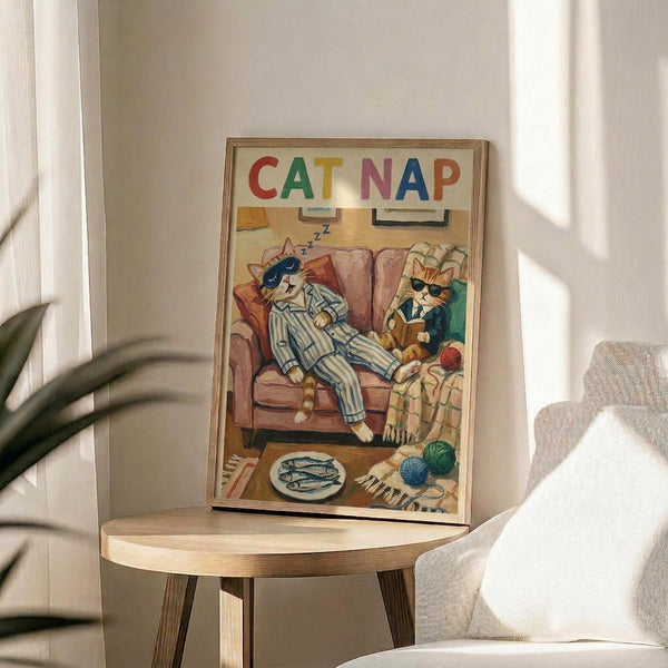 May include: Framed poster with colorful "CAT NAP" text showing two anthropomorphic cats relaxing on a pink armchair - one sleeping in striped pajamas, another reading while wearing sunglasses. Scene includes fish on plate, yarn ball, and cozy living room details. Print displayed on round wooden side table next to beige upholstered chair with plant shadows casting across white walls in bright, naturally lit room.