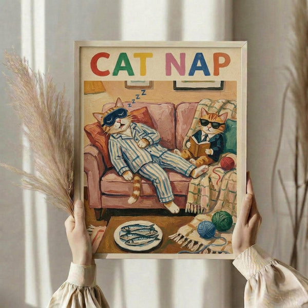 May include: Hands holding a framed vintage-style illustration showing two orange cats on a brown couch - one sleeping in blue striped pajamas, another wearing sunglasses and reading. Colorful "CAT NAP" lettering at top. Scene includes yarn balls, fish plate, and cozy living room details. Dried pampas grass visible beside the light wood frame against cream curtains.