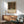 May include: Framed Gustav Klimt poster featuring a lush garden scene with vibrant sunflowers, red poppies, and dense wildflowers in rich greens and earth tones, displayed above a mid-century modern wooden sideboard. Light green upholstered armchair with wooden frame sits nearby, alongside a white ceramic table lamp and small round coffee table, all bathed in natural window light creating geometric shadows on white walls.